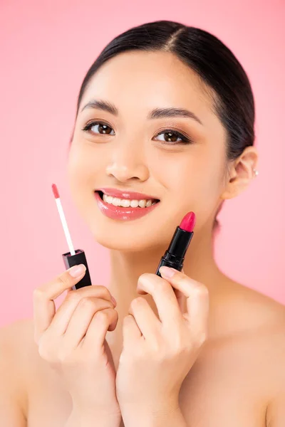 Junge asiatische Frau blickt in die Kamera, während sie Lipgloss und Lippenstift isoliert auf rosa hält — Stockfoto