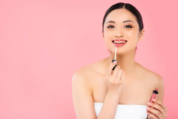 Brünette asiatische Frau Anwendung Lipgloss und Blick auf Kamera isoliert auf rosa — Stockfoto