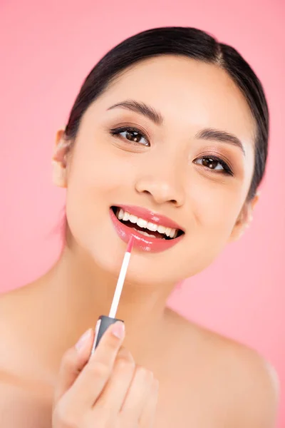 Brünette asiatische Frau schaut in die Kamera, während sie Lipgloss isoliert auf rosa anwendet — Stockfoto