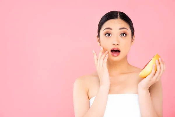 Conmocionada mujer asiática tocando la cara mientras sostiene la mitad de pomelo aislado en rosa — Stock Photo