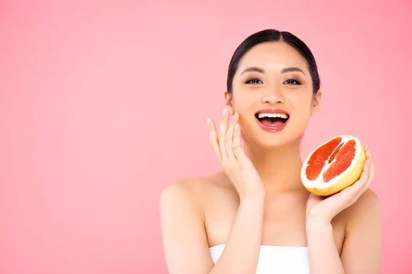 Збуджена азіатська жінка тримає половину соковитого грейпфрута, торкаючись обличчя ізольовано на рожевому — стокове фото