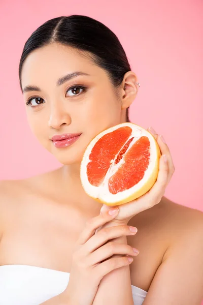 Young asian woman holding half of ripe grapefruit while looking at camera isolated on pink — Stock Photo