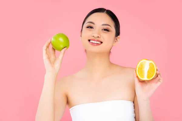 Молода азіатка тримає зелене яблуко і половину стиглого апельсина ізольовано на рожевому — стокове фото