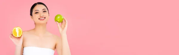 Panoramic concept of asian woman holding whole apple and half of juicy orange isolated on pink — Stock Photo