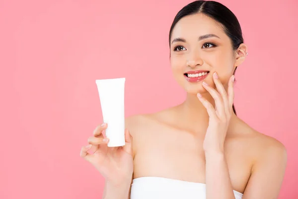 Bruna donna asiatica in possesso di tubo di crema cosmetica, toccando il viso e guardando lontano isolato sul rosa — Foto stock