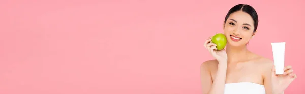 Horizontal concept of asian woman holding cosmetic cream and whole green apple isolated on pink — Stock Photo