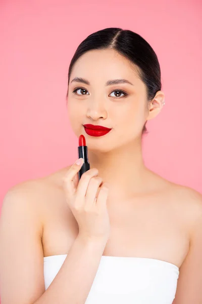 Junge asiatische Frau blickt in die Kamera, während sie roten Lippenstift in Gesichtsnähe hält, isoliert auf rosa — Stockfoto