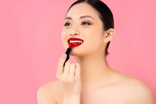 Nackte asiatische Frau schaut weg, während sie roten Lippenstift isoliert auf rosa aufträgt — Stockfoto