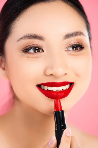 Nahaufnahme einer jungen asiatischen Frau, die roten Lippenstift aufträgt, während sie isoliert von rosa wegschaut — Stockfoto