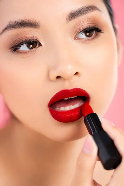 Primer plano vista de joven asiático mujer aplicando rojo lápiz labial aislado en rosa - foto de stock