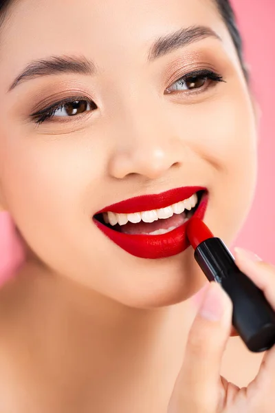 Primer plano vista de asiático mujer aplicando rojo lápiz labial aislado en rosa - foto de stock