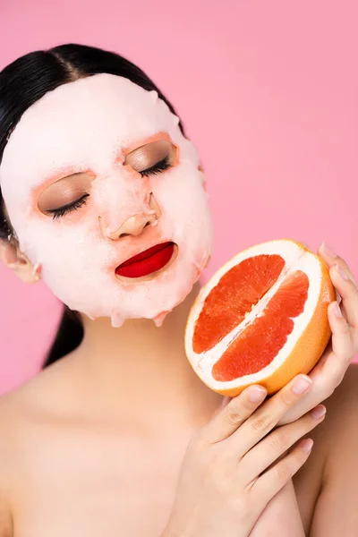 Morena asiática mujer en la cara máscara celebración de la mitad de maduro pomelo con los ojos cerrados aislado en rosa - foto de stock