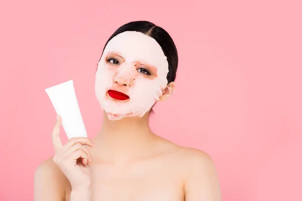 Joven asiático mujer en la cara máscara celebración tubo de cosmética crema aislado en rosa - foto de stock