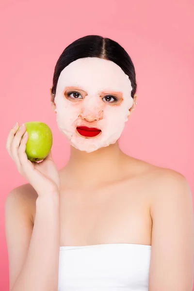 Jeune asiatique femme dans visage masque tenant mûr vert pomme tout en regardant caméra isolé sur rose — Photo de stock