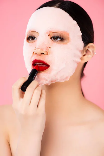 Оголена азіатська жінка в масці для обличчя наносить червону помаду ізольовано на рожевий — стокове фото