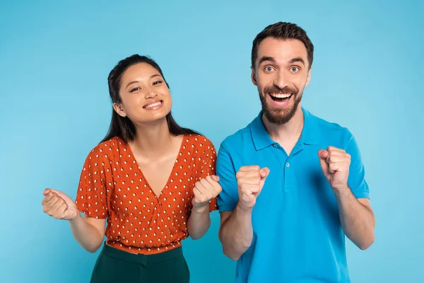 Excité homme en polo t-shirt et asiatique femme en chemisier rouge montrant victoire geste sur bleu — Photo de stock