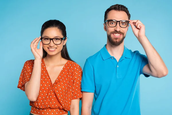 Jeune couple interracial regardant la caméra tout en touchant les lunettes isolées sur bleu — Photo de stock