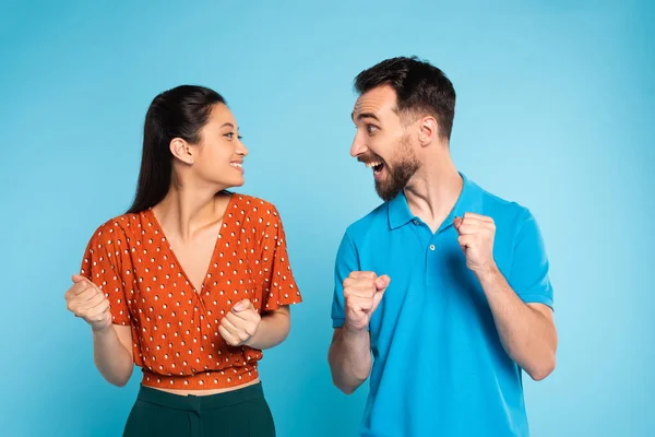 Aufgeregter Mann im Polo-T-Shirt und asiatische Frau in roter Bluse schauen einander an, während sie Siegergeste auf blau zeigen — Stockfoto