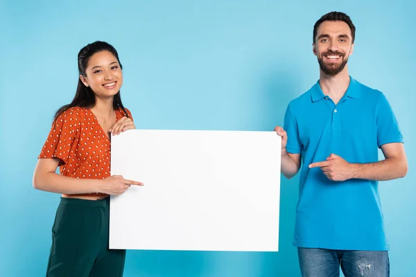 Asiatin in roter Bluse und Mann im Polo-T-Shirt zeigen mit den Fingern auf leeres Plakat auf blauem Grund — Stockfoto