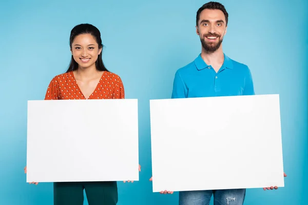 Young interracial couple looking at camera while holding blank placards on blue — Stock Photo