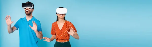 Panoramic concept of excited man in polo t-shirt and woman in red blouse gesturing with hands while using vr headsets on blue — Stock Photo