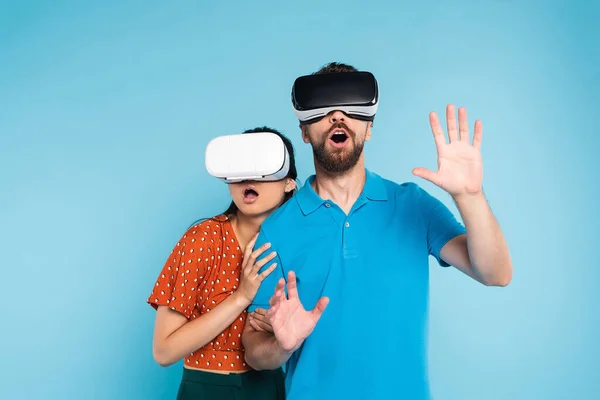 Scared woman in red blouse touching excited man in polo t-shirt while using vr headsets together isolated on blue — Stock Photo