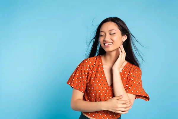 Elegant asian woman in red blouse touching neck while posing with closed eyes on blue — Stock Photo