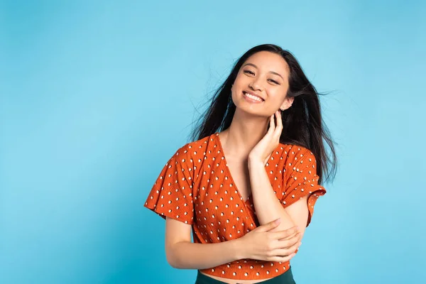 Asian woman in red stylish blouse touching neck and looking at camera on blue — Stock Photo