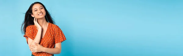 Horizontal concept of elegant asian woman in red blouse looking at camera while touching neck on blue — Stock Photo