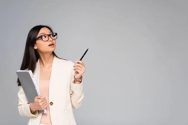 Surpris asiatique femme d'affaires en blanc blazer regardant loin et pointant avec stylo isolé sur gris — Photo de stock