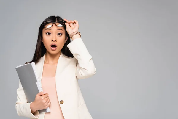 Überraschte asiatische Geschäftsfrau im weißen Blazer, die eine Brille berührt, während sie mit offenem Mund in die Kamera schaut, isoliert auf grau — Stockfoto