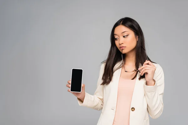 Thoughtful asian businesswoman in blazer holding eyeglasses while showing smartphone with blank screen isolated on grey — Stock Photo