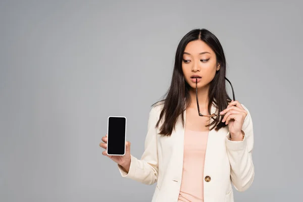 Pensativa mujer de negocios asiática mostrando teléfono inteligente con pantalla en blanco mientras sostiene las gafas aisladas en gris - foto de stock