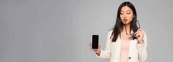 Horizontal image of thoughtful asian businesswoman holding eyeglasses while showing smartphone with blank screen isolated on grey — Stock Photo