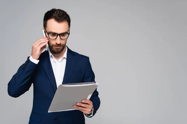 Homme d'affaires sérieux en blazer bleu et lunettes regardant des documents tout en parlant sur smartphone isolé sur gris — Photo de stock