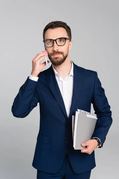 Seriöser Geschäftsmann mit Ordner, der wegschaut, während er auf dem Smartphone isoliert auf grau spricht — Stockfoto