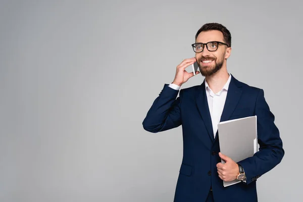 Бородатый бизнесмен в синем пиджаке и очках смотрит в сторону, разговаривая на смартфоне, изолированном на сером — стоковое фото
