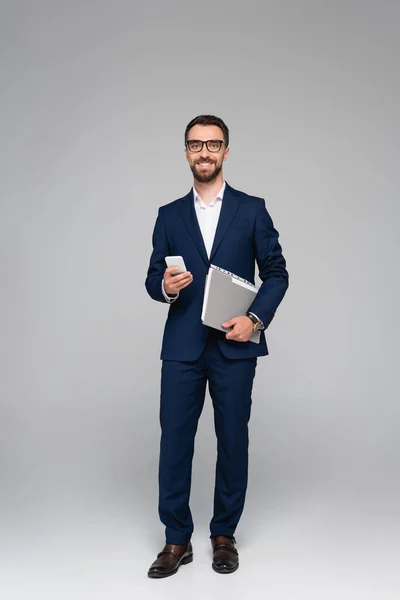 Bearded businessman in blue suit and glasses holding smartphone and folder on grey — Stock Photo