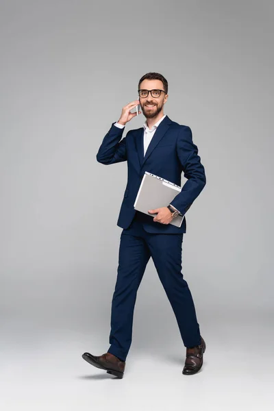 Businessman in blue suit talking on smartphone and holding folder while walking on grey — Stock Photo