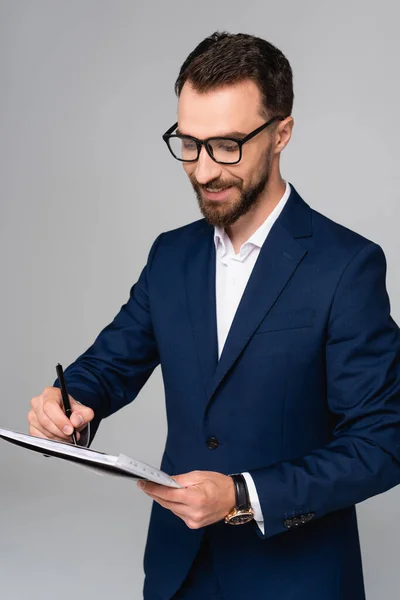 Joven hombre de negocios en blazer azul y anteojos escrito en documento aislado en gris - foto de stock