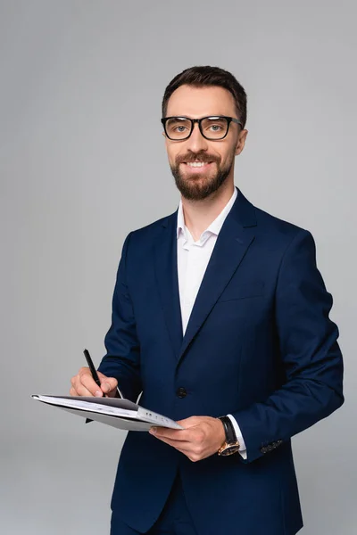 Bearded businessman in blue blazer and eyeglasses holding folder and pen isolated on grey — Stock Photo