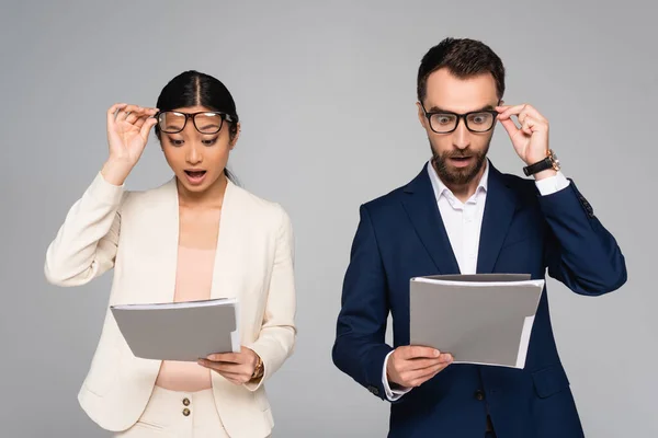 Surprised interracial business partners looking at documents and touching eyeglasses isolated on grey — Stock Photo