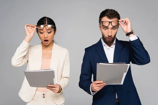 Impactado interracial pareja de socios de negocios tocando gafas mientras mira carpetas aisladas en gris - foto de stock
