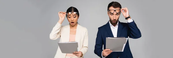Website-Kopfzeile eines überraschten interrassischen Paares von Geschäftskollegen, die eine Brille berühren, während sie Ordner isoliert auf grau anschauen — Stockfoto