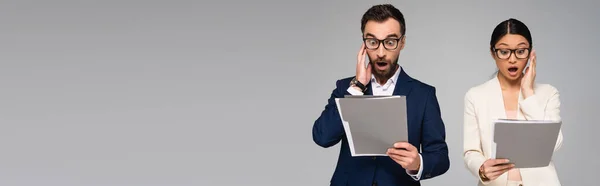 Panoramic crop of shocked interracial businesspeople holding hands near faces while looking at folders isolated on grey — Stock Photo