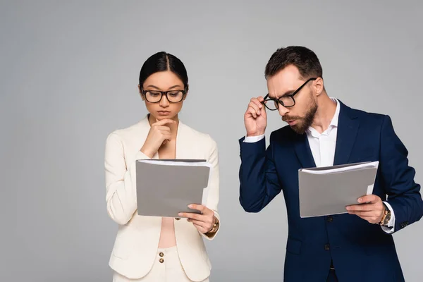 Thoughtful asian businesswoman touching chin while looking at documents near colleague isolated on grey — Stock Photo