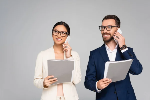 Couple interracial de jeunes partenaires commerciaux tenant des dossiers et parlant sur des téléphones mobiles isolés sur gris — Photo de stock