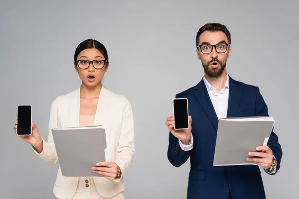 Geschockte Geschäftspartner halten Ordner in die Höhe, während sie Mobiltelefone mit leerem Bildschirm in grau zeigen — Stockfoto