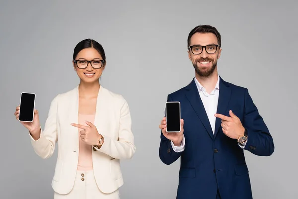 Pareja interracial de jóvenes socios de negocios apuntando a teléfonos inteligentes con pantalla en blanco aislado en gris - foto de stock