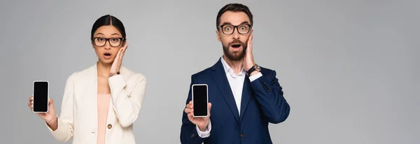 Schockierte Geschäftsleute, die Gesichter berühren, während sie Smartphones mit leerem Bildschirm zeigen, isoliert auf grauem, horizontalem Konzept — Stockfoto